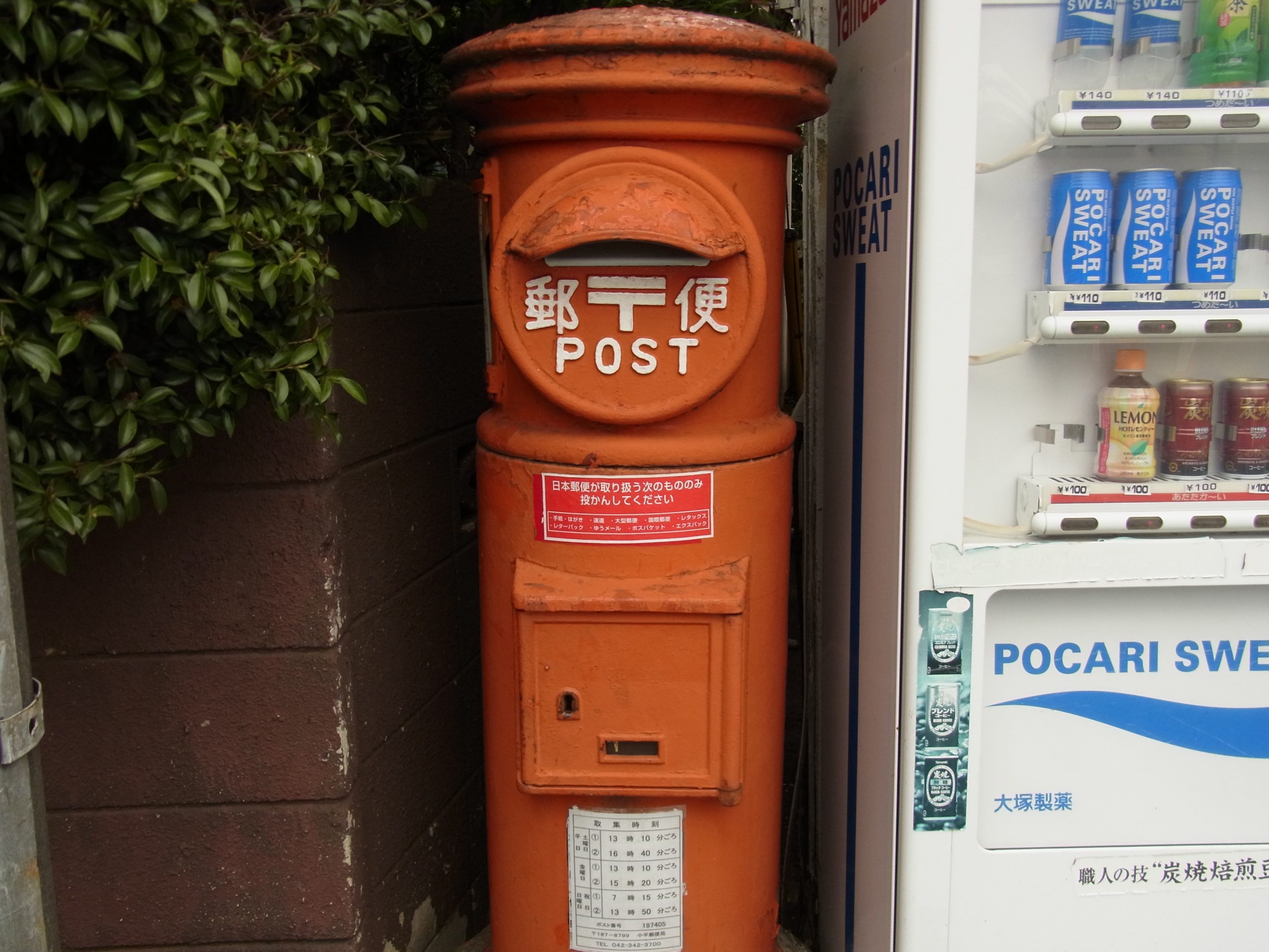 自販機の隣に佇むオレンジ色のレトロな郵便ポストです。